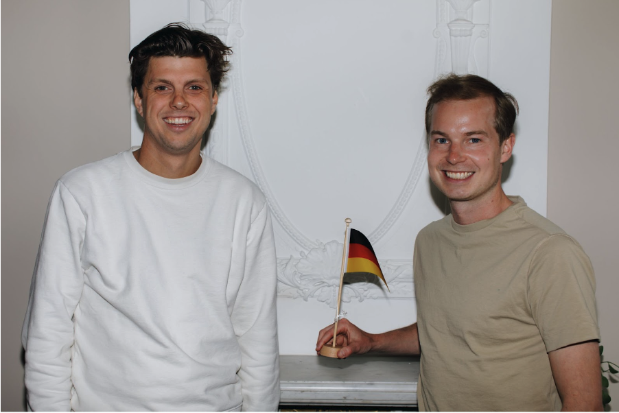 Twee oprichters van Brenger, Wisse Koedam en Derk van der Have, staan lachend naast elkaar en houden een kleine Duitse vlag vast.