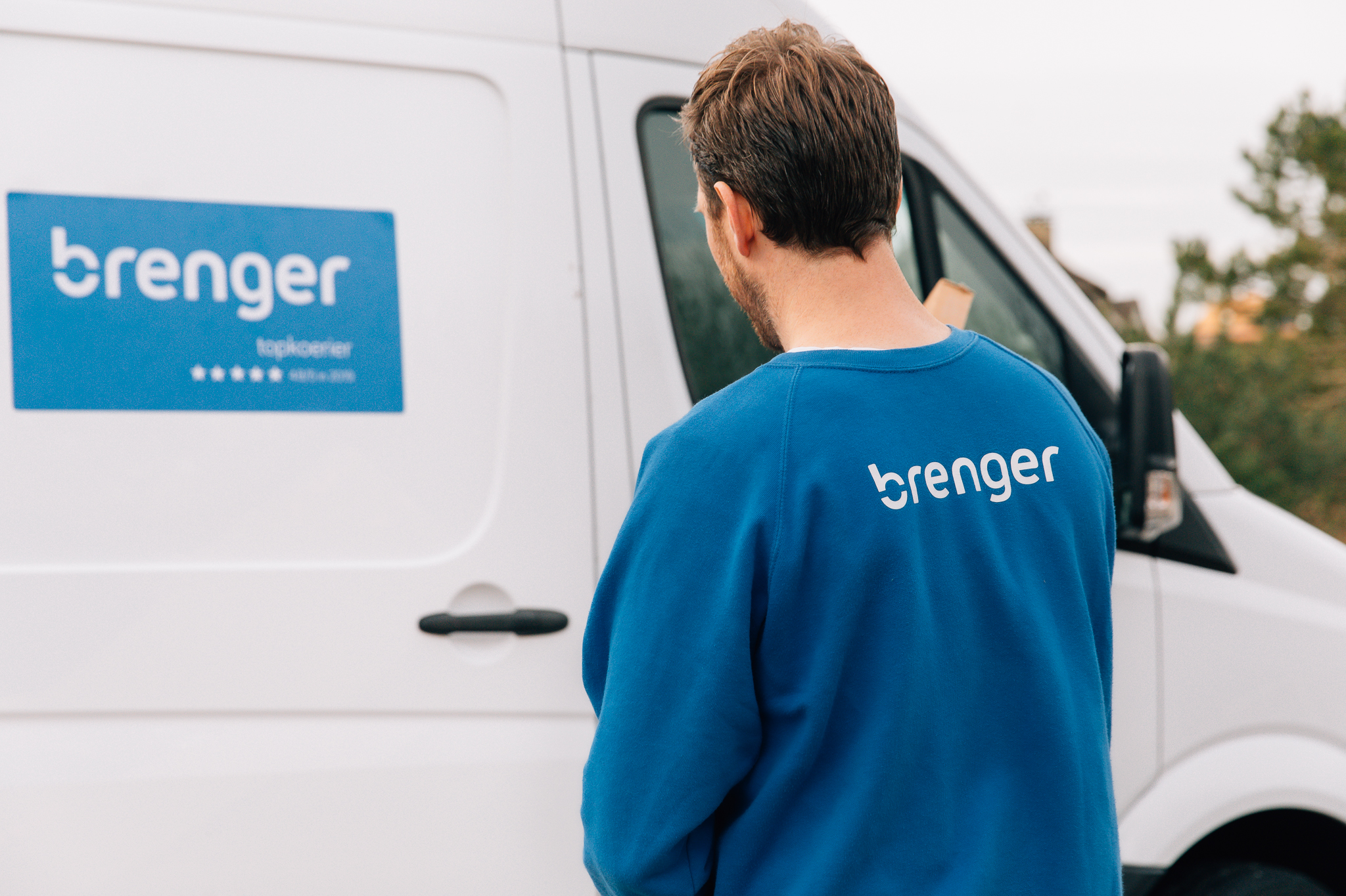 Een Brenger-koerier in een blauw uniform staat naast een witte bestelbus met Brenger-logo, klaar om een pakketje te bezorgen.