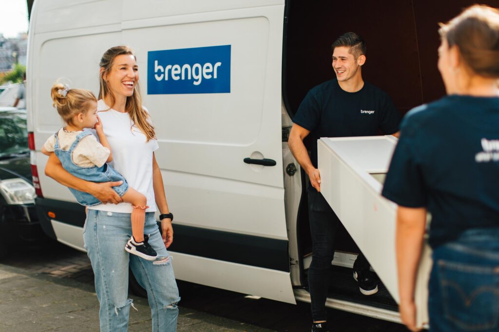 Een vrouw met een jong kind op haar arm staat lachend naast een bestelwagen van Brenger, terwijl een medewerker van Brenger een wit meubelstuk uit de bestelwagen tilt.